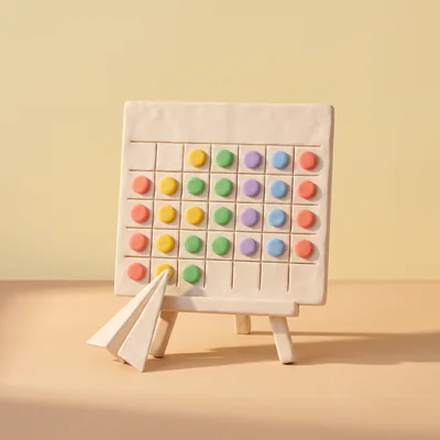 Un calendrier en pâte sur un chevalet, ses cases remplies de pastilles de couleur, un avion en papier à son pied.