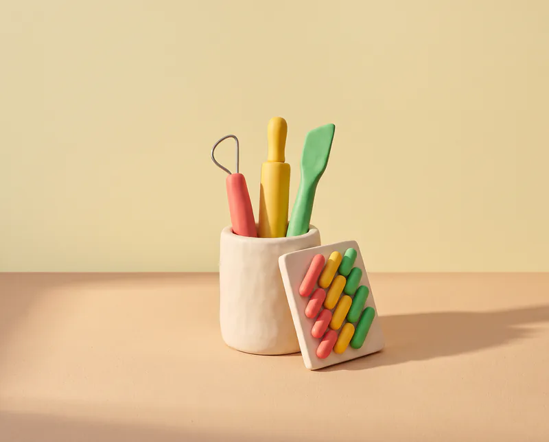 Trois outils de modelage aux manches rouge, jaune et vert rangés dans un pot en pâte, un petit tableau de pilules appuyé contre le pot : l’atelier d’Ethanolle.